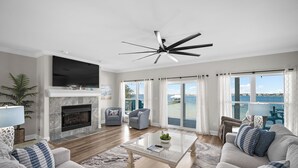 A living room with a fireplace, a ceiling fan, a large TV, and a view of the sea. - Spacious Perdido Key Retreat: Sand Volleyball, Dock, Gameroom & Beach (Pensacola)