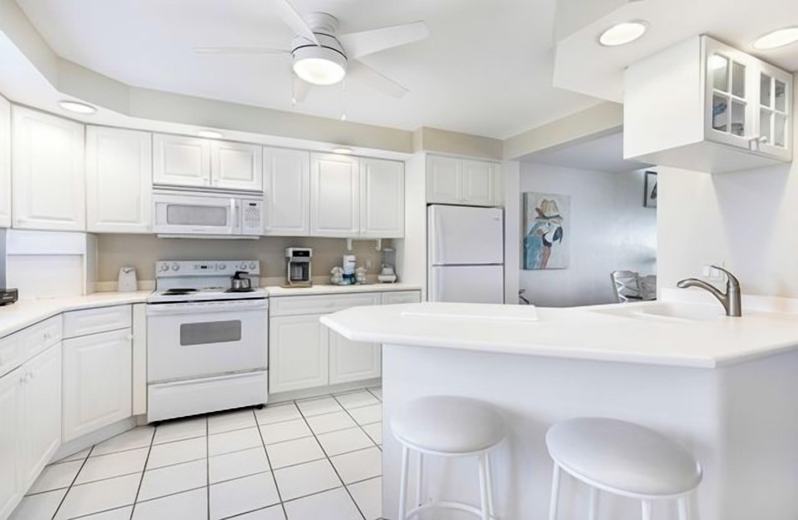 Une cuisine moderne avec des meubles blancs, un îlot central doté de tabourets, un four à micro-ondes, une cuisinière, un réfrigérateur et un ventilateur de plafond.