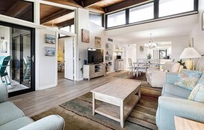 Een ruime woonkamer met een houten salontafel, een bank en een televisie. Er is een eethoek met een kroonluchter en een keuken op de achtergrond.