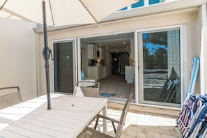 Uma área de pátio externa com mesa e cadeiras, um grande guarda-sol e uma porta de vidro deslizante que dá para uma cozinha e sala de estar.