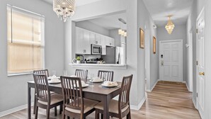 A dining area with a wooden table and chairs, a kitchen in the background, and a hallway leading to a door.