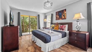 A bedroom with a large bed, a wooden dresser, a nightstand with a lamp, a painting on the wall, and a view of the outdoors through a sliding door.