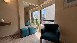 Ein modernes Wohnzimmer mit einem großen Fenster, einem Topf mit Pflanze, türkisfarbenen Hocker und einem dunkel-türkisfarbenen Sessel.