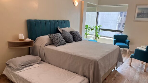 Ein modernes Schlafzimmer mit einem großen Bett, einem Schlafsofa, einem Nachttisch, einem Sessel und einem Fenster mit Blick auf Gebäude.