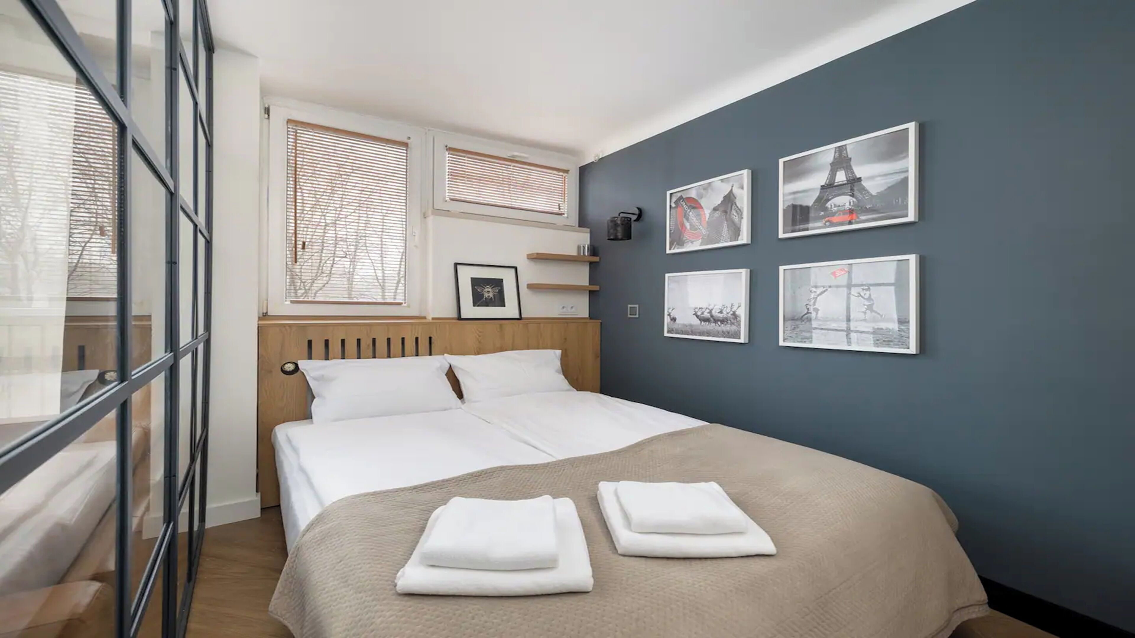 Una cama bien hecha con sábanas blancas, una manta color beige y dos toallas dobladas sobre la cama. Hay cuatro cuadros en la pared, incluyendo una fotografía de la Torre Eiffel.