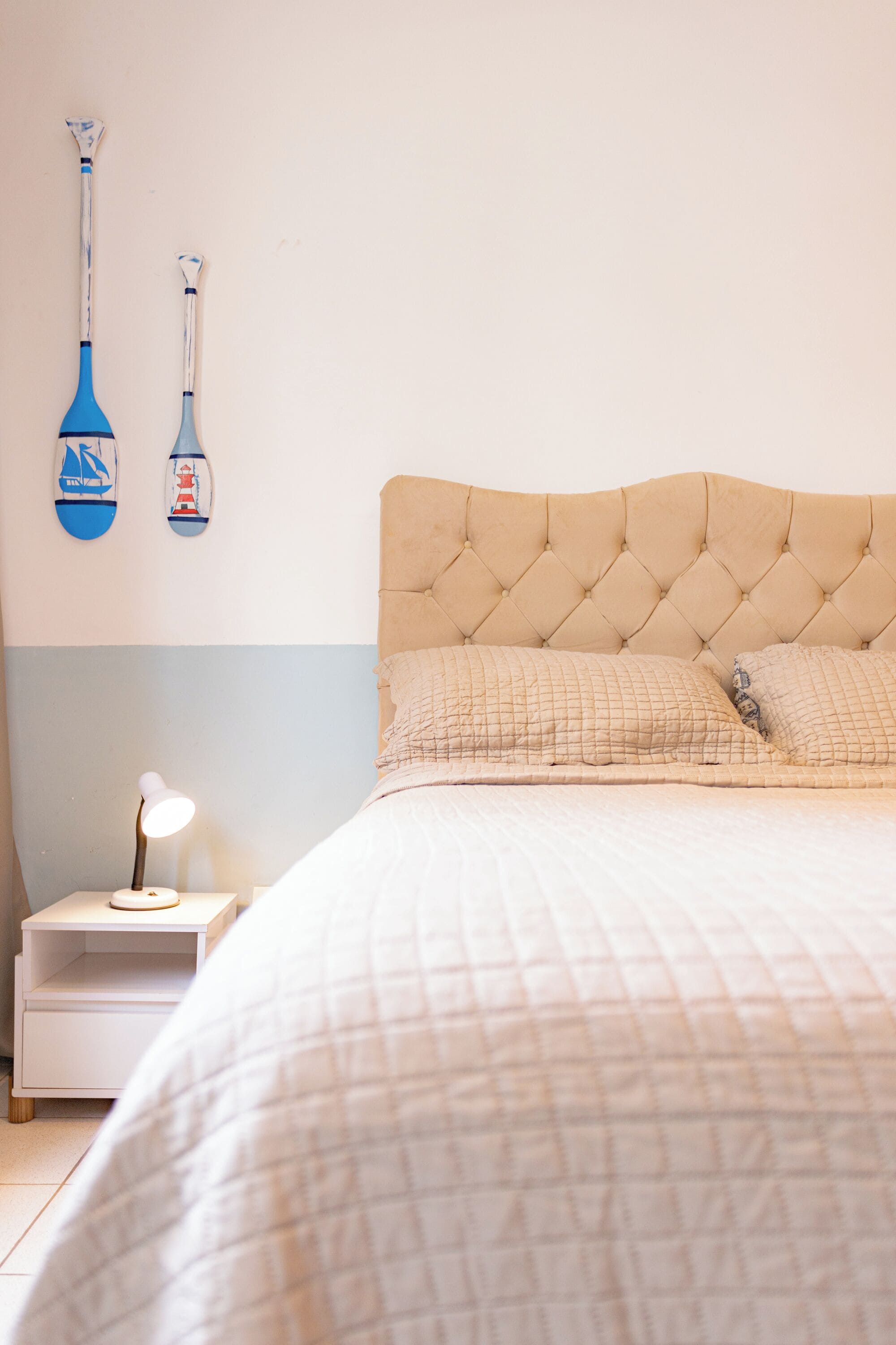 A neatly made bed with a quilted headboard and pillows, a bedside table with a lamp, and two wall-mounted items.