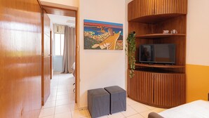 A hotel room with a wooden wardrobe, a television on a wooden shelf, a painting on the wall, and a small grey ottoman.