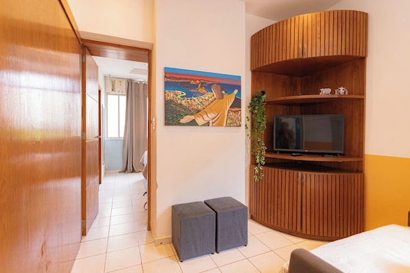 A hotel room with a wooden wardrobe, a television on a wooden shelf, a painting on the wall, and a small grey ottoman.