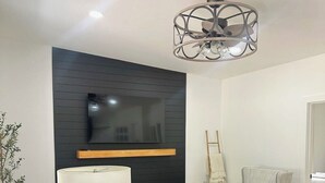 Una sala de estar moderna con un televisor de pantalla plana montado en una pared negra, un sofá color beige, un reposapiés azul, una mesita de madera con una lámpara y una lámpara decorativa en el techo.