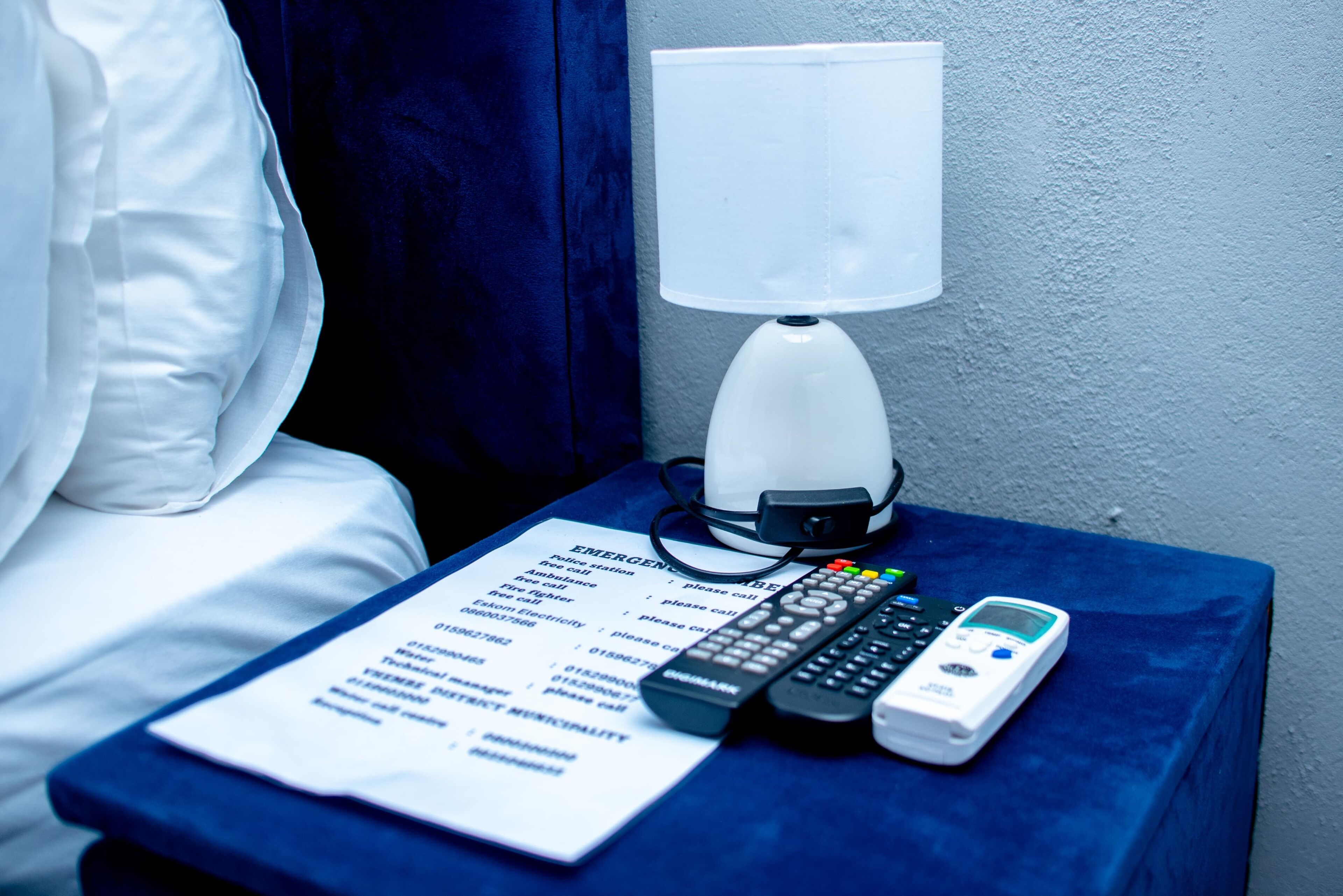 Een hotelkamer met een bed, een nachtkastje met een lamp, een afstandsbediening, een mobiele telefoon en een lijst met noodcontacten.