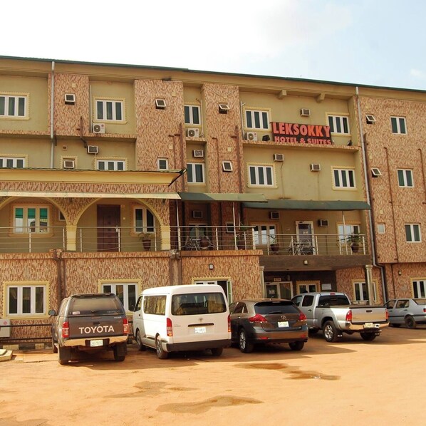 A multi-story building with a sign for 'LEKSOKKY HOTEL & SUITES'. There are several parked vehicles in front. - Lekosokky  Hotel and Suites (Ijoko)