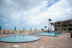 A pool area with lounge chairs and umbrellas, a beach view, and a building with balconies.