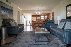 A living room with a sofa, coffee table, and a rug. There is a kitchen in the background with cabinets and a dining area.