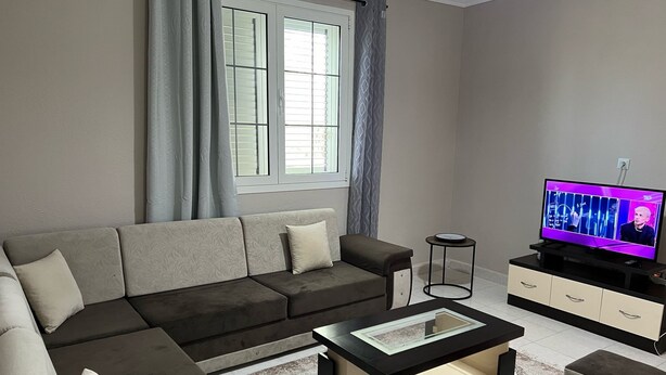 A modern living room with a sectional sofa, a coffee table, ottomans, a TV, and a small side table. - Elodi’s Villa (Fushkruje)