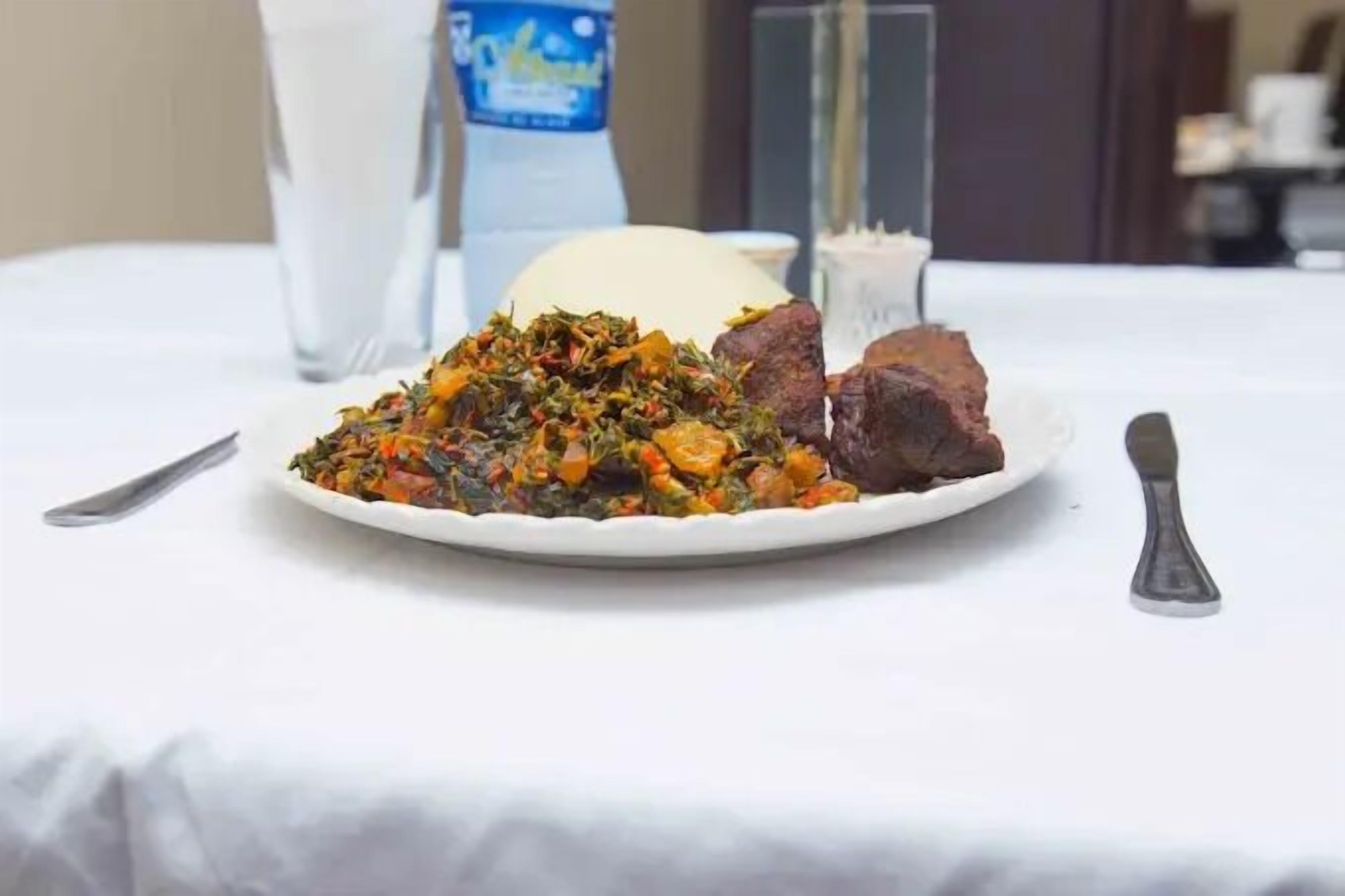 Un plato de comida con guarnición de verduras, un trozo de carne, un vaso de agua y una botella de agua sobre una mesa.