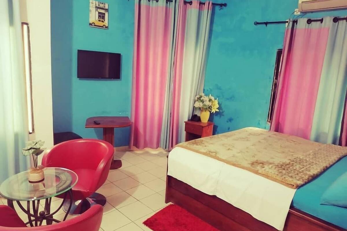 Ein Schlafzimmer mit Bett, Fernseher, kleinem Tisch, Stuhl und Vorhängen.