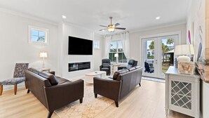 A modern living room with a flat-screen TV, leather sofas, a fireplace, and a balcony with a view of palm trees. - Sugar Shores at Summer Salt (Orange Beach)