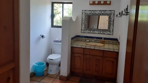 Een kleine badkamer met toilet, wastafel en spiegel. Boven het toilet is een raam en op de vloer staat een blauwe emmer.