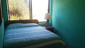 Een slaapkamer met een bed, nachtkastje, lamp en raam met uitzicht op bomen.