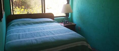 Un dormitorio con cama, mesita de noche, lámpara y ventana con vista a árboles.