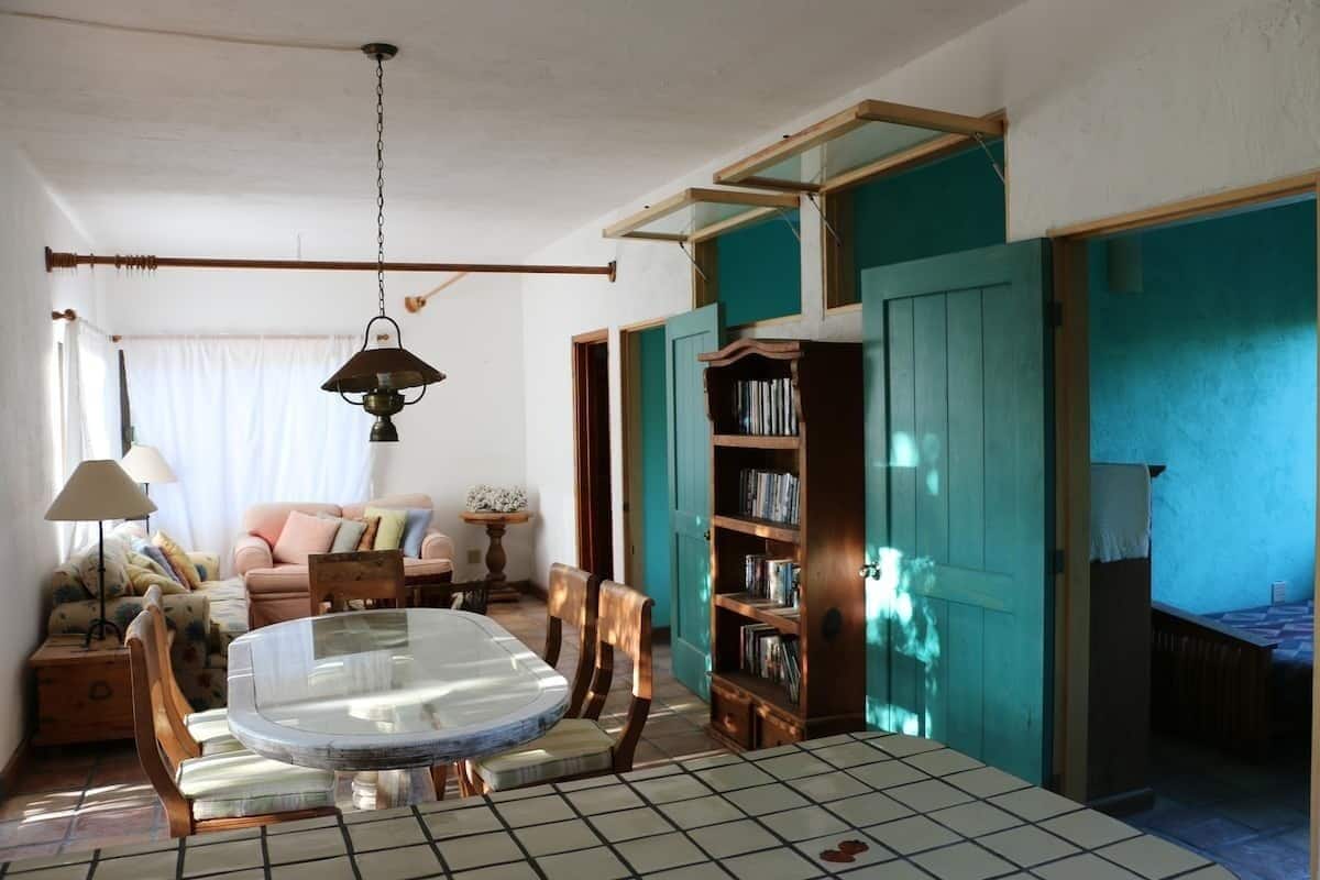 A cozy living room with a round glass table, wooden chairs, a bookshelf, and a teal door.
