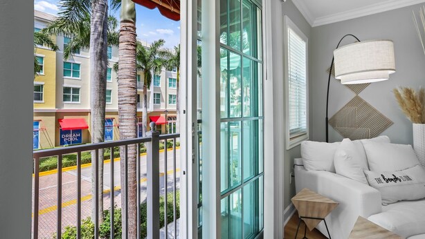 A balcony with a view of palm trees and buildings, a white sofa with a lamp, and a small side table.