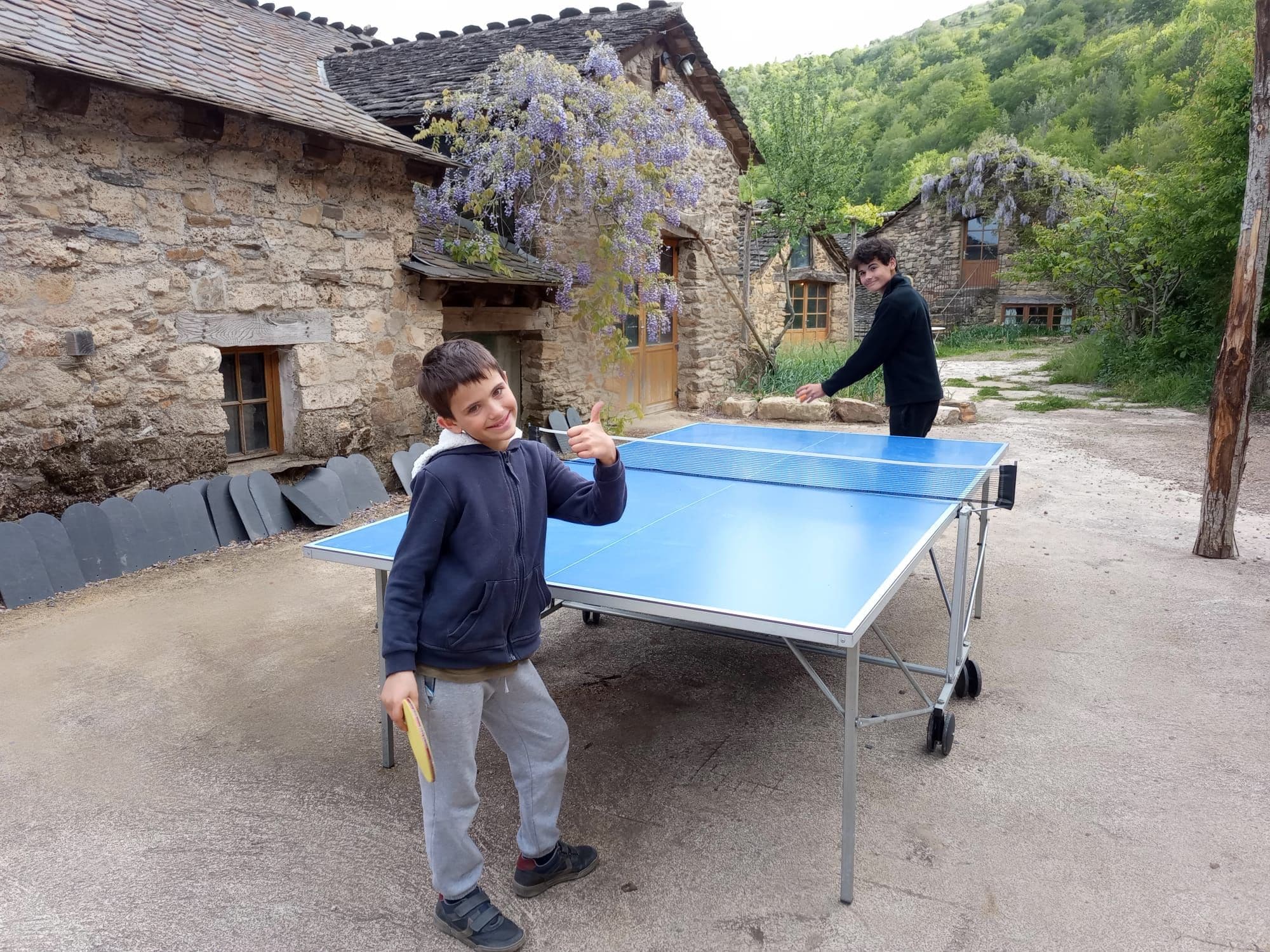 Two individuals playing table tennis outdoors in front of a stone building with a traditional sloped roof.