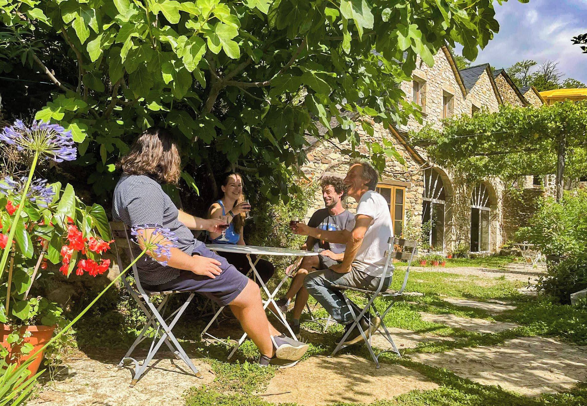 Un groupe de personnes est assis à une table à l’extérieur, dans un jardin, avec des bâtiments en pierre en arrière-plan.