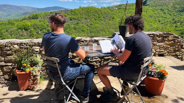Dos personas sentadas en una mesa con vistas a colinas verdes y un cielo despejado.