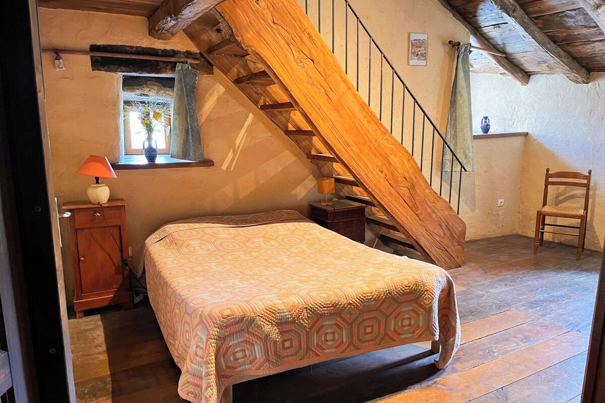 Une chambre avec un escalier en bois, un lit recouvert d’une couvre-lit à motifs, une table de chevet, une lampe et une fenêtre avec un rideau.