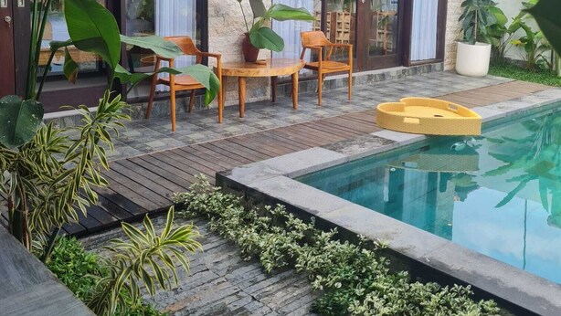 Un área de piscina moderna al aire libre con cubierta de madera, dos sillas de madera y una plataforma flotante amarilla.