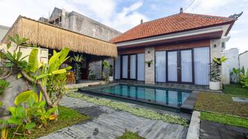 A modern house with a swimming pool, a thatched-roof structure, and a garden with tropical plants.