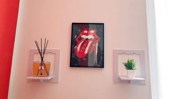 A wall with a framed artwork of red lips, two small shelves with a diffuser and a plant, and a fire extinguisher sign. - Rosa Homes THE RED (Genova)