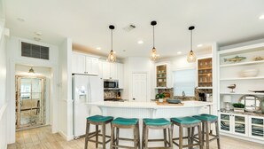 A kitchen with white cabinets, a central island with bar stools, pendant lighting, and a built-in shelving unit with decorative items. - Pet Friendly, Easy Walk to Beach, Game Room, Beach Toys, Gorgeous Community (Galveston)
