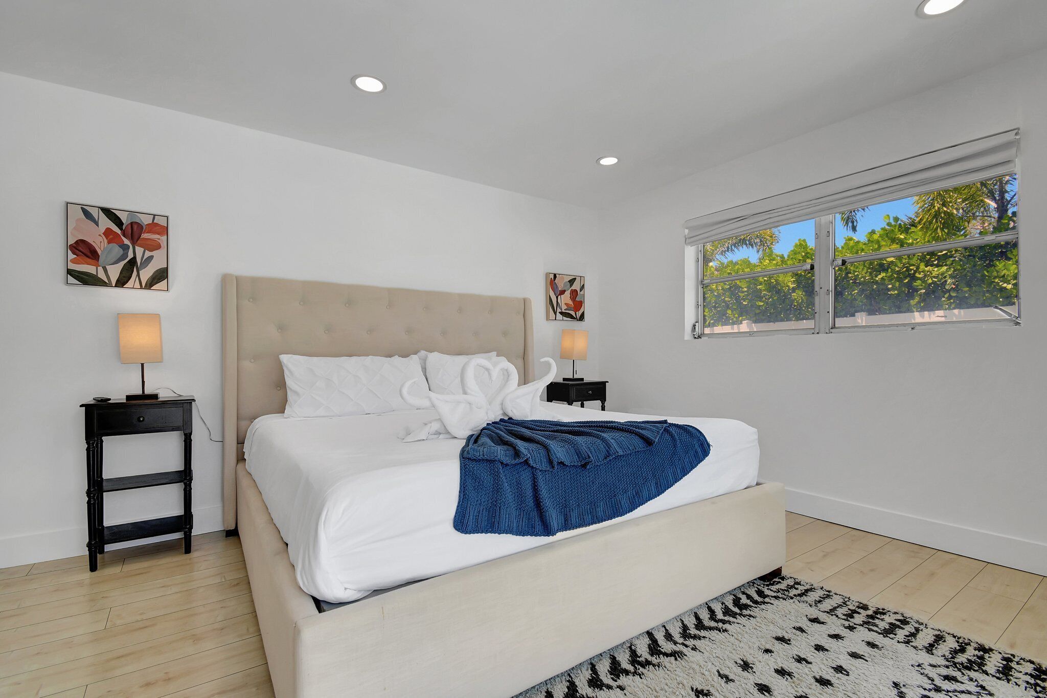 A bedroom with a bed, two bedside tables, a window with a view of trees, and two wall-mounted lamps.