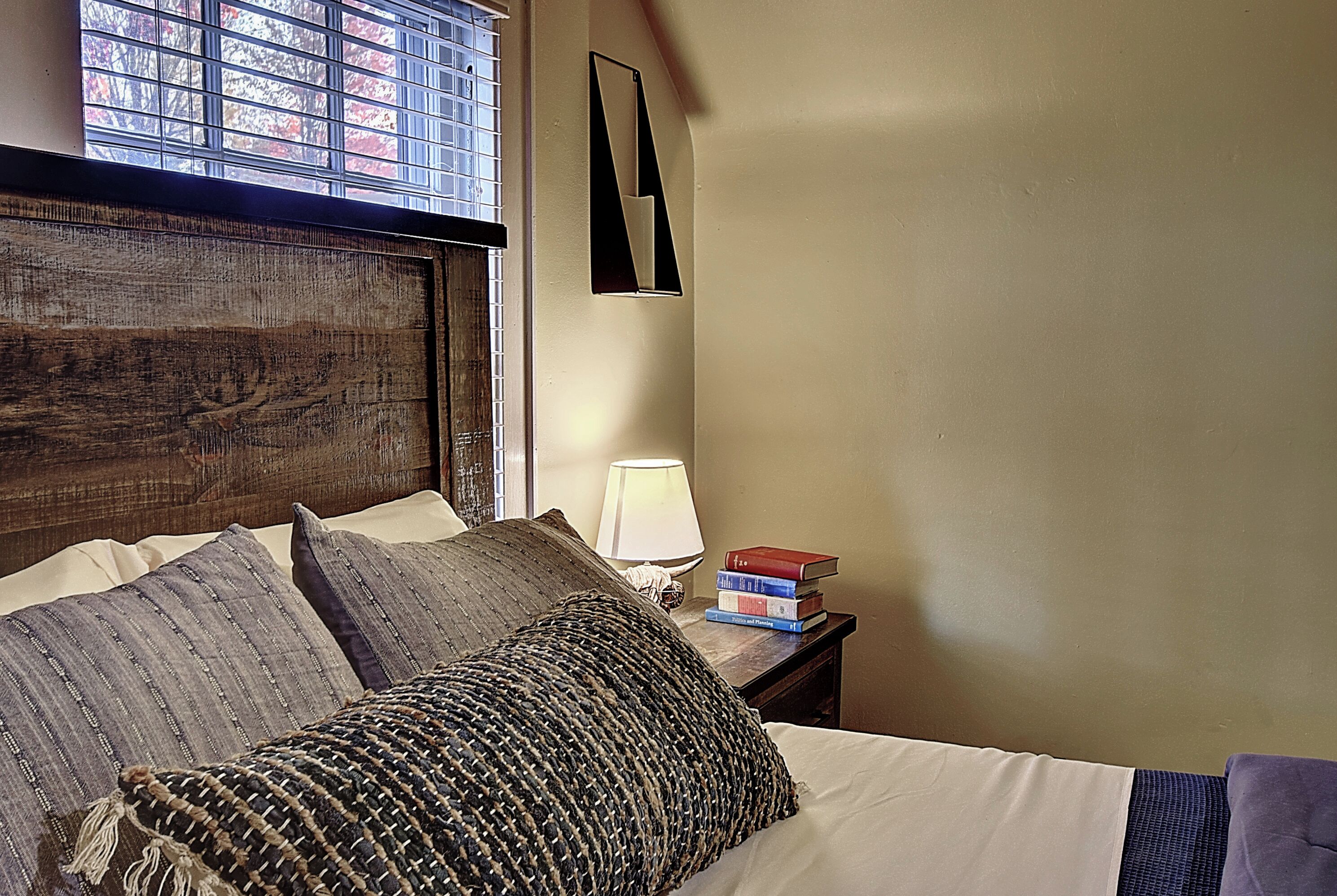 Una cama bien hecha con varias almohadas, una lámpara de noche y una pila de libros en la mesita.
