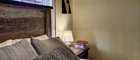 Una cama bien hecha con varias almohadas, una lámpara de noche y una pila de libros en la mesita.