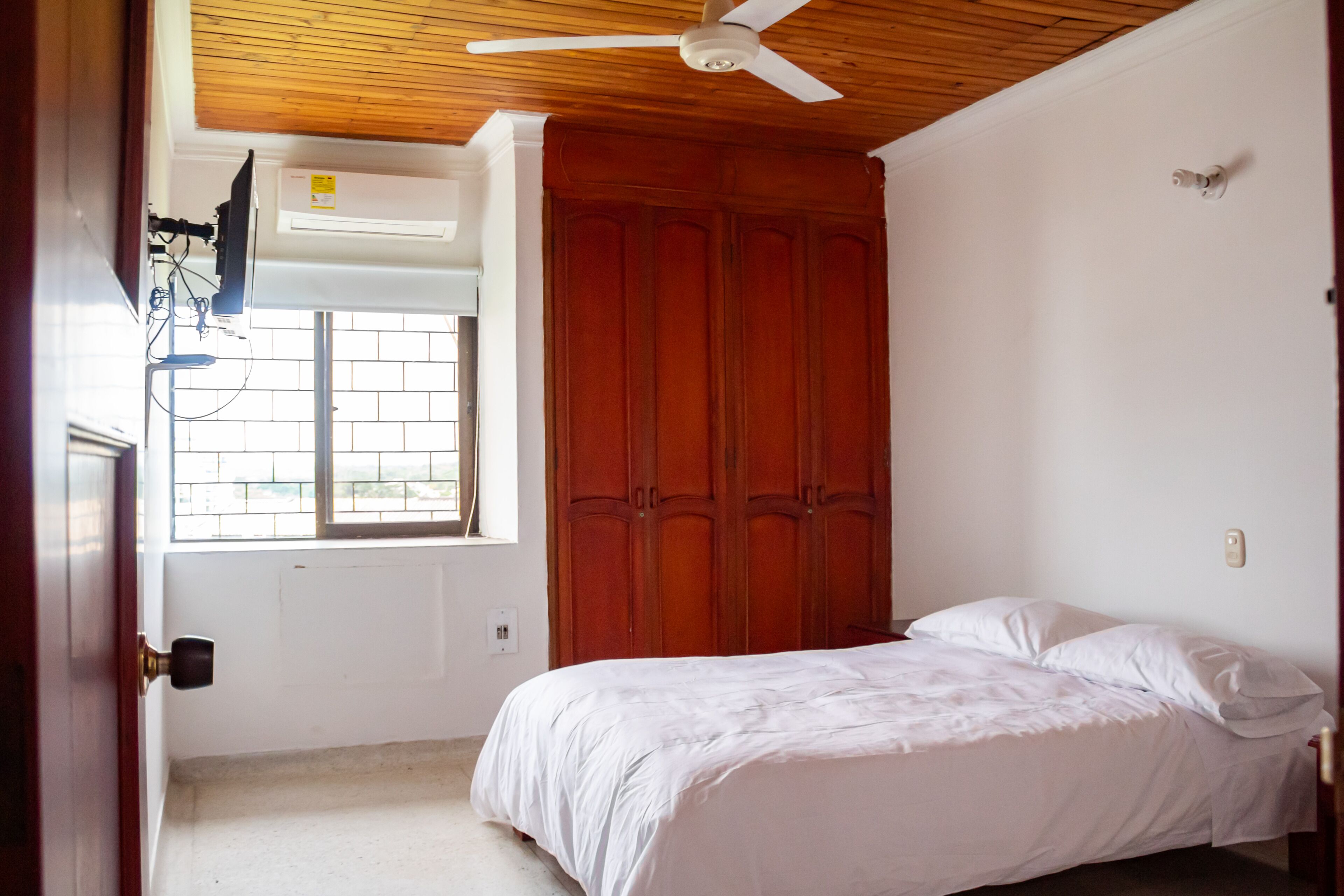 A bedroom with a wooden ceiling, a window with a view, a bed, and a wardrobe.