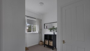 Un couloir avec une porte blanche, une fenêtre ornée de rideaux blancs, une console noire sur laquelle sont posés des paniers, et une plante.