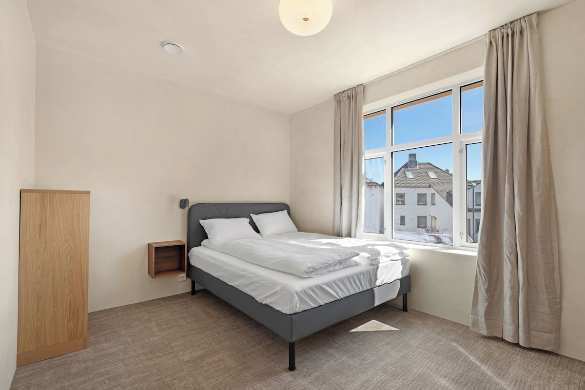 Une chambre à coucher moderne avec un lit, une table de chevet, une armoire en bois et une grande fenêtre avec des rideaux.