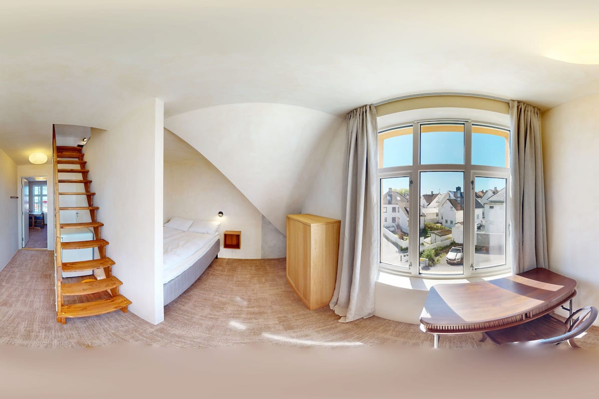 Une pièce spacieuse avec un plafond voûté, un escalier en bois, un lit, une petite table et une grande fenêtre avec des rideaux.