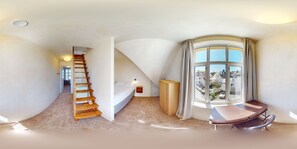 Une pièce spacieuse avec un plafond voûté, un escalier en bois, un lit, une petite table et une grande fenêtre avec des rideaux.