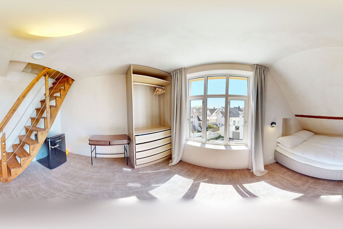 Une chambre avec un plafond incurvé, un lit, une fenêtre avec des rideaux, un escalier en bois et une petite table.