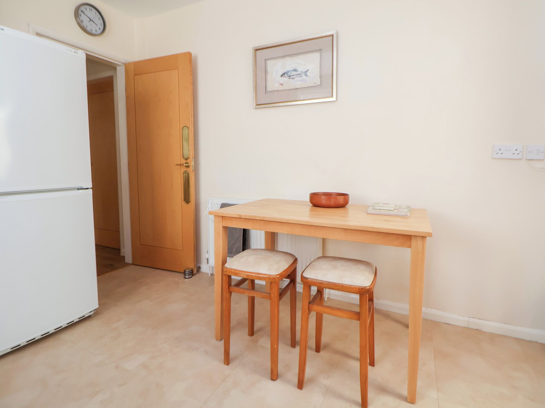 Eine kleine Küche mit einem weißen Kühlschrank, einem Holztisch mit zwei Stühlen, einem gerahmten Bild an der Wand und einer Uhr über der Tür.