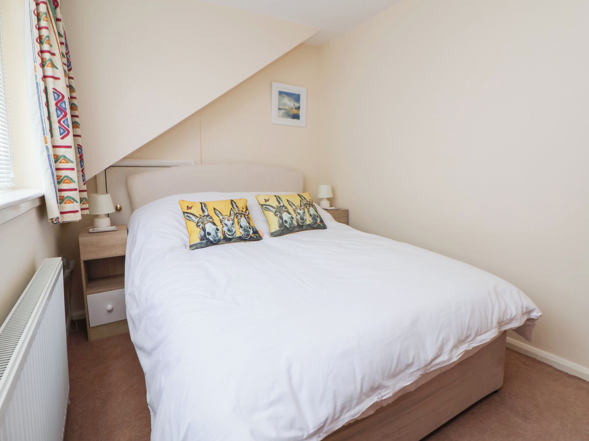A bedroom with a bed, two bedside tables with lamps, a radiator, a window with curtains, and a picture on the wall.