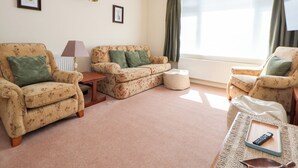 A living room with floral-patterned furniture, a lamp, a remote control on a tray, and a window with curtains. - Bryn Bach (Colwyn Bay)
