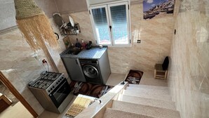 A kitchen with a gas stove, oven, and washing machine. There is a window with a view of boats and a decorative wall hanging.