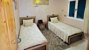 A room with two single beds, a wooden wardrobe, a small table with a laptop, a decorative ceiling light, and a wall with Arabic calligraphy.