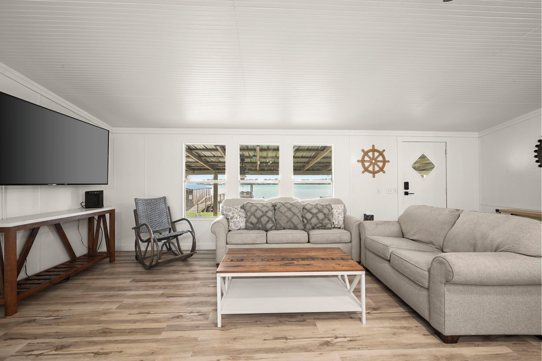 A modern living room with a large flat-screen TV, two sofas, a coffee table, and a view of the outdoors through large windows.
