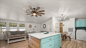 Un vaste espace de vie comprenant un salon avec une table basse en bois, un canapé et une cuisine dotée de meubles bleu clair et d’un lave-linge blanc.